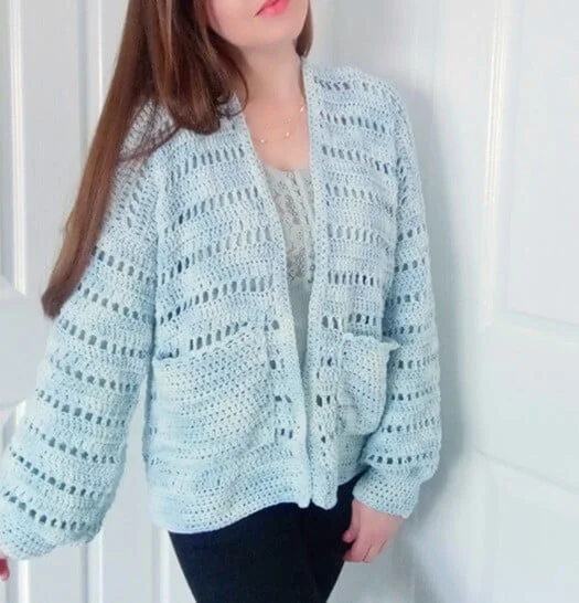 Woman leaning against a wall wearing a pale blue crochet summer cardigan with long sleeves, horizontal openwork rows, and two front pockets over a lace-trimmed top and dark pants.