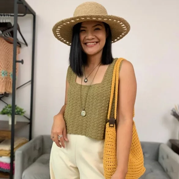 An olive coloured crochet tank top is styled on a woman. She has accessorised with a straw hat and orange shoulder bag