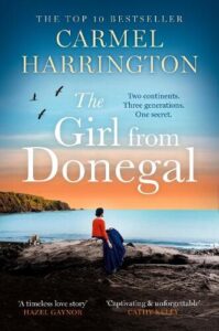 book cover for The Girl from Donegal by Carmel Harrington. A dark haired woman wearing a red jumper is sat on a sea wall facing a bay