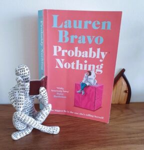 A paperback copy of Probably Nothing by Lauren Bravo is on a shelf with a small statue showing a person sitting crossed legged and reading a book