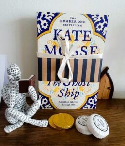 A paperback copy of The Ghost Ship by Kate Mosse is on a shef with an ornament of a person reading a book