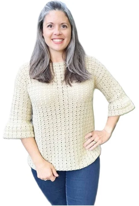 Woman wearing a light beige crochet sweater with subtle bell sleeves and a vertically textured stitch pattern. The top has a relaxed fit and is paired with dark blue jeans, shown against a white background with the model smiling confidently.
