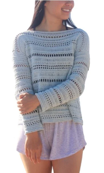 Woman smiling while wearing a light blue crochet sweater with horizontal openwork rows and a relaxed fit. The sweater has long sleeves and a boat neckline, paired with soft lavender shorts, and is styled in a breezy, beach-ready look against a white background.