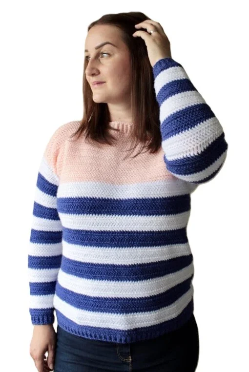 Woman wearing a crew neck crochet sweater with horizontal stripes in soft pink, white, and navy blue. The sweater has a fitted shape with long sleeves and ribbed cuffs and hem, styled casually with dark jeans against a white background.