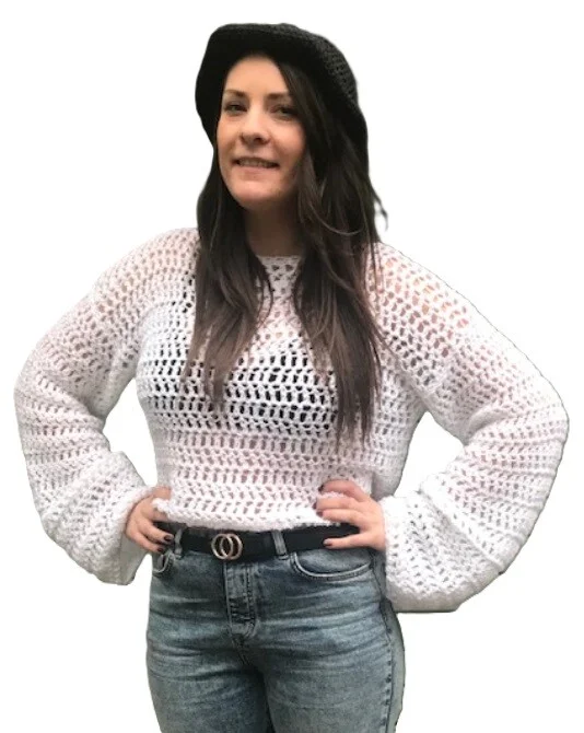 Woman wearing a white crochet crop top with a lacy open stitch pattern, long balloon sleeves, and a round neckline. She styles it with high-waisted jeans, a black belt, and a black hat, posing with hands on hips against a white background.