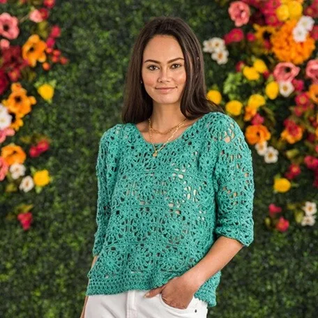 Woman wearing a teal, lacy crochet sweater made of floral motifs, standing in front of a vibrant green hedge wall decorated with colorful flowers. She has long brown hair, a relaxed smile, and is dressed casually with one hand in the pocket of her white pants.