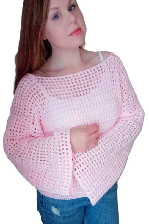 Woman wearing a soft pink crochet mesh sweater with wide bell sleeves and a loose, off-the-shoulder fit. The sweater features an open stitch pattern layered over a matching tank top, paired with blue jeans, against a white background.