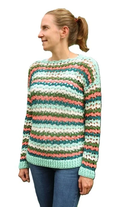 Woman wearing a colorful crochet sweater with horizontal stripes in shades of mint green, coral, teal, and olive. The sweater features a lacy stitch pattern with a ribbed hem and cuffs, styled casually with blue jeans against a white background.