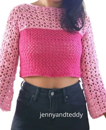 Woman wearing a two-tone pink crochet crop sweater with a solid stitch pattern on the body and lacy openwork on the sleeves and upper chest. The sweater has a boat neckline and long sleeves, styled with high-waisted black jeans;