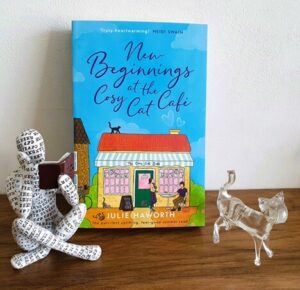 Paperback copy of New Beginnings at the Cosy Cat Cafe by Julie Haworth is resting on a shelf with a glass cat ornament and a person sitting crossed legged reading a book (an ornament)