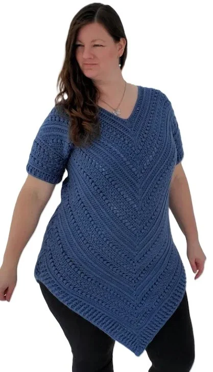 Woman wearing a deep blue crochet tunic sweater with short sleeves and a textured chevron stitch pattern. The tunic features a V-neckline and a pointed hemline that creates an elongated silhouette, styled with black leggings against a white background.