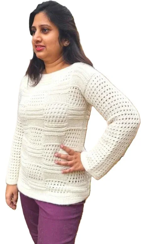 Woman wearing an off-white Tunisian crochet sweater featuring a mix of textured blocks and lacy stitch panels. The sweater has long sleeves and a classic crew neck, styled with plum-colored pants and shown against a white background.