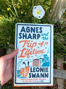 A hand is holding up a hardcover copy of Agnes Sharp and the Trip of a Lifetime by Leonie Swann. In the background is greenery and a white flower