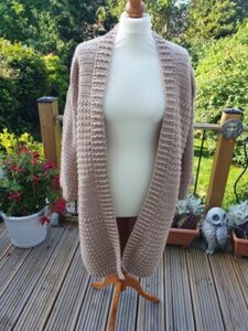 A long cardigan in a beige colour is styled on a mannequin. The mannequin is on a decking with trees in the background