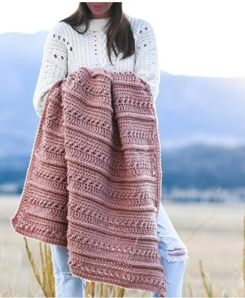 A woman is standing in a field with mountains behind her.  She is holding a blanket