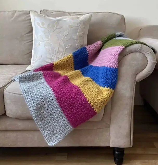 A blanket crocheted in stripes of grey, maroon, mustard, blue and pink is draped over the arm of a sofa