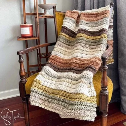 A chunky blanket crocheted in stripes of different shades of brown is draped over a chair