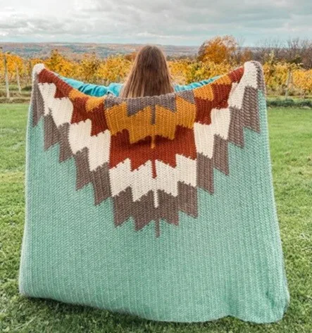 A woman is standing facing away from the viewer in a field.  She is holding up a blanket to show the tapestry design