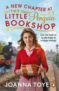 Book cover for A New Chapter at the Little Penguin Bookshop by Joanna Toye. A woman in a pink gingham dress and red cardigan is carrying books as she walks along the railway platform