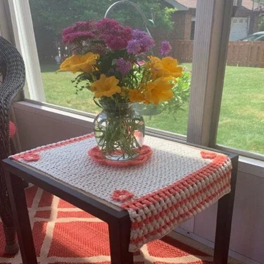 A square table is decorated with a table runner with a crocheted flower centre piece and a flower at each corner