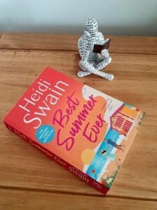 Paperback of Best Summer Ever by Heidi Swain is on a wooden surface. An ornament of a person sitting crossed legged reading a book is next to the paperback