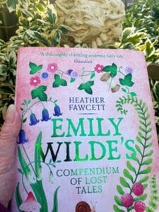 A person holds the book "Emily Wilde’s Compendium of Lost Tales" by Heather Fawcett. The book cover is pink with green leaves, bluebells, mushrooms, and illustrated flowers, along with a snail near the bottom. In the background, there’s lush green foliage and a stone garden ornament of a face peeking through the leaves.