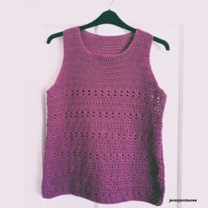 A purple crochet summer vest with a sleeveless, scoop-neck design hanging on a black hanger against a pale pink door. The summer top features alternating rows of solid and open lacy stitches, creating a breathable, textured look. The bottom right corner includes the text “jerasjamboree.”