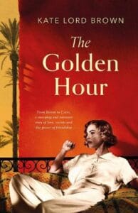 Cover of The Golden Hour by Kate Lord Brown. The design features a sepia-toned image of a glamorous woman reclining on an ornate couch, set against a bold gradient background of red fading into golden yellow. A single palm tree is silhouetted on the left. Text reads: “From Biarritz to Cairo, a sweeping and intimate story of love, secrets and the power of friendship.”