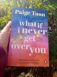 A person holds up the book What If I Never Get Over You by Paige Toon outdoors in natural sunlight, with greenery and purple flowers in the background. The book cover features a colorful gradient of blue to orange with sparkler illustrations and glitter-like accents. The tagline reads: "Three days to fall in love. Six years to try to forget."