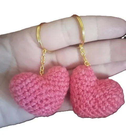 A pair of plush, red-pink crochet heart keychains with gold chains and keyrings, displayed in the palm of a hand. The hearts are stuffed for a three-dimensional shape, giving them a soft, squeezable appearance.