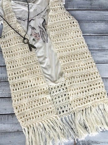 Cream-colored crochet vest laid flat over a printed tee, featuring open stitch patterns and long fringe along the hem. The vest has a vintage, boho-inspired feel, styled with a leather necklace and key charm for a rustic vibe.