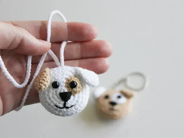 A close-up of a handmade crochet dog face keychain in white and beige, featuring black safety eyes, a black nose, and a stitched smile, held in a person's hand. Another similar dog keychain is blurred in the background, showing its metal keyring attachment.
