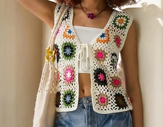 Woman wearing a sleeveless crochet vest made from colorful granny squares bordered in white, paired with a white crop top and denim shorts. The vest has a casual, boho vibe, with a drawstring tie at the front and a handmade flower necklace to match.