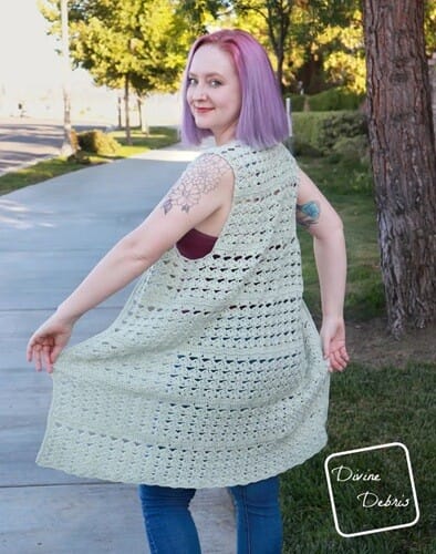 Woman with pastel purple hair modeling a sleeveless, light sage green crochet vest with an open lacy stitch pattern. She’s twirling slightly on a sunny sidewalk, smiling over her shoulder