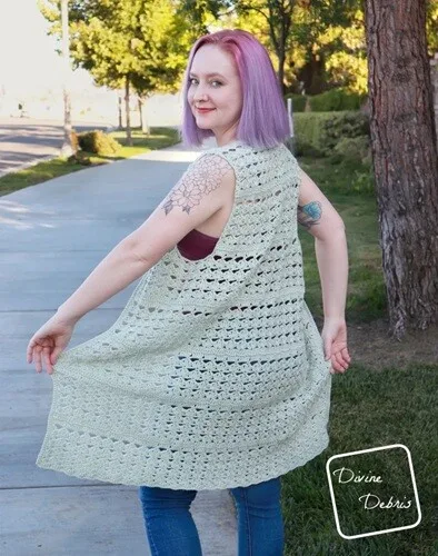 Woman with pastel purple hair modeling a sleeveless, light sage green crochet vest with an open lacy stitch pattern. She’s twirling slightly on a sunny sidewalk, smiling over her shoulder