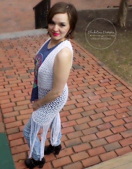 Woman wearing a long, white crochet vest with a mesh-style stitch and long fringe, styled over a graphic tee and jeans. She’s standing on a red brick walkway, glancing over her shoulder, with a soft smile