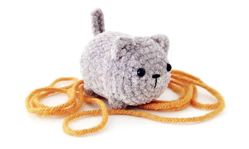 A plush, no-sew crochet cat made with soft, light gray yarn, featuring black safety eyes, a tiny nose, and a curved tail. The cat is shown playfully positioned among loops of mustard-yellow yarn on a white background.