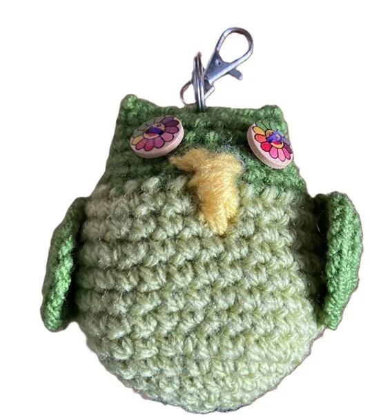 A handmade green crochet owl keychain with outstretched wings, a yellow beak, and colorful wooden button eyes featuring flower patterns. The keychain has a silver clasp attached at the top for easy clipping to bags or keys.