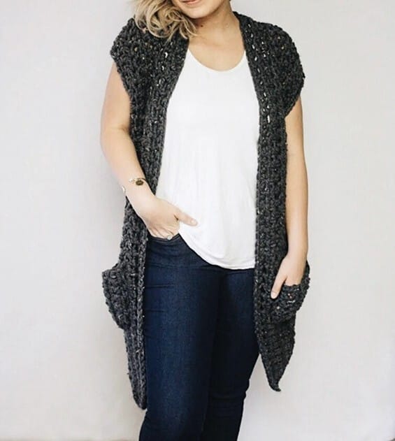 Woman wearing a chunky, dark gray crochet vest with cap sleeves and deep front pockets, styled over a white tee and skinny jeans. The vest features a soft, textured stitch pattern and drapes casually at the sides for a relaxed, comfy look.