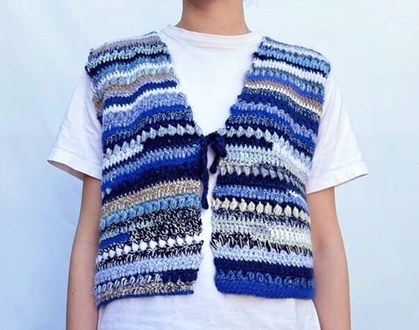 Close-up of a person wearing a handmade crochet vest with horizontal stripes in shades of blue, cream, black, and tan. The vest features a mix of textured and open stitches, tied at the front with a simple cord, layered over a plain white t-shirt.