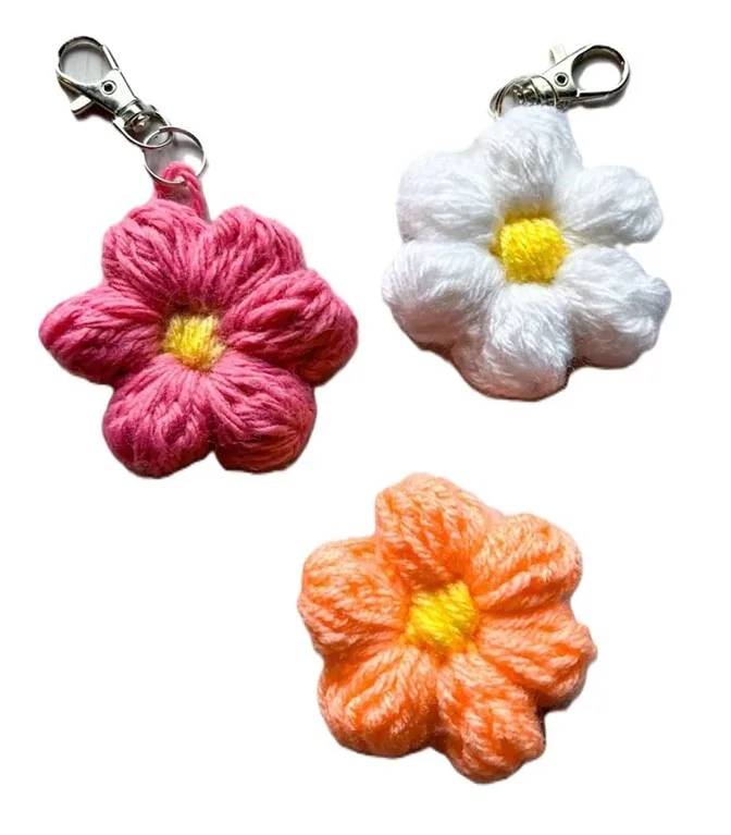 Three crochet flower keychains in pink, white, and orange with bright yellow centers, two featuring metal lobster clasps for easy attachment. The flowers have a puffy, textured look with six petals each, designed to resemble cheerful blooms made from soft yarn.