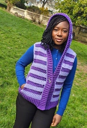 Woman wearing a handmade crochet hooded vest with bold purple and lavender stripes, styled over a blue long-sleeve shirt and black pants. The vest has a thick ribbed trim and a relaxed fit, perfect for layering on cool days in the park.