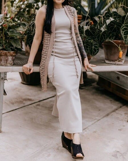 Woman wearing a light beige crochet vest with simple, open stitches over a matching sleeveless maxi dress, seated on a bench surrounded by potted plants. The vest has long drawstrings and adds a breezy, handmade touch to the elegant, minimalist look.