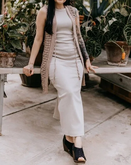 Woman wearing a light beige crochet vest with simple, open stitches over a matching sleeveless maxi dress, seated on a bench surrounded by potted plants. The vest has long drawstrings and adds a breezy, handmade touch to the elegant, minimalist look.