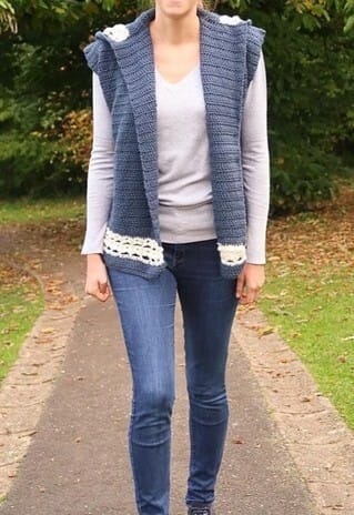 Woman walking down a park path wearing a blue crochet hooded vest with cream trim along the hem. The vest is styled over a light gray long-sleeve shirt and jeans, featuring a simple textured stitch and cozy hood for added warmth.