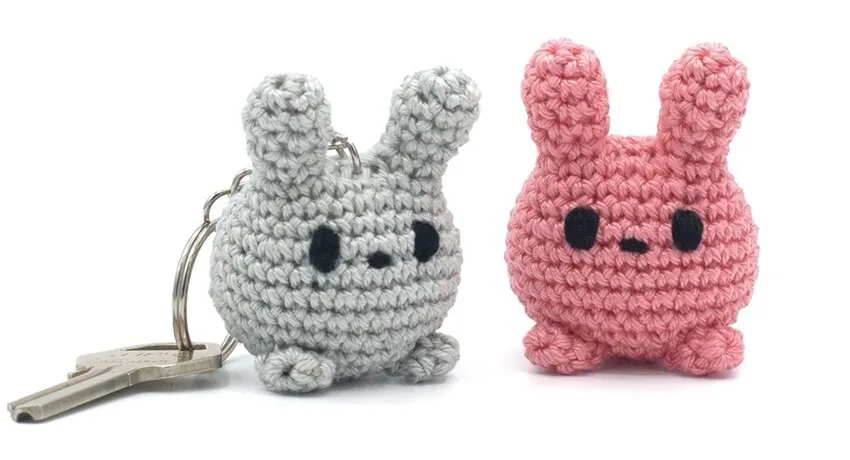 Two round crochet bunny figures, one in gray with an attached keyring and one in pink, both with black embroidered eyes, long upright ears, and small feet. The gray bunny is shown with a metal key, highlighting its use as a keychain.