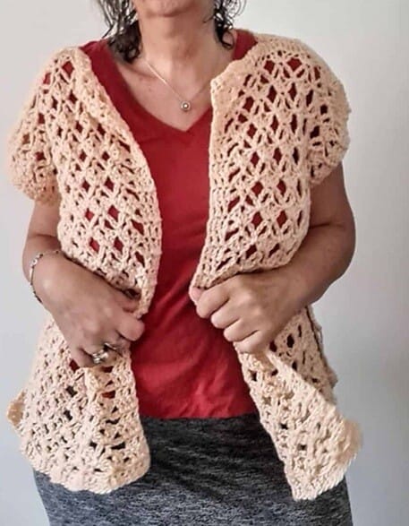Woman wearing a short-sleeved, open-front crochet vest with a lacy diamond stitch pattern in a soft peach color. The vest is styled casually over a red V-neck shirt and a heather gray skirt, showing off its airy texture and drape.