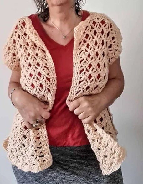 Woman wearing a short-sleeved, open-front crochet vest with a lacy diamond stitch pattern in a soft peach color. The vest is styled casually over a red V-neck shirt and a heather gray skirt, showing off its airy texture and drape.