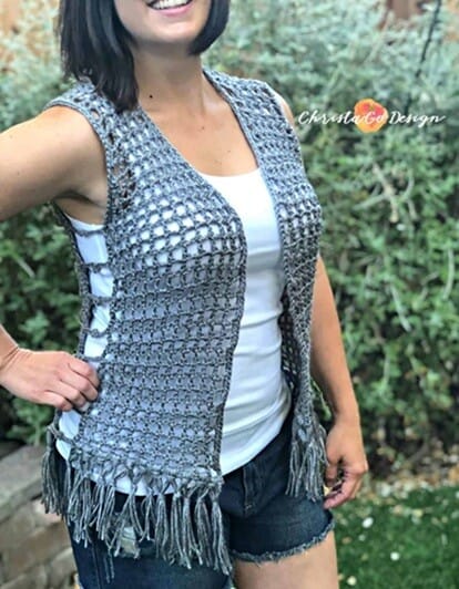 Woman wearing a sleeveless gray crochet vest with open mesh stitches and fringe along the bottom edge. The vest features side ties and is styled over a white tank top and denim shorts, perfect for a casual, boho-inspired summer look.