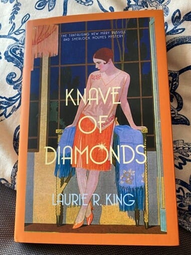 Hardback copy of Knave of Diamonds by Laurie R. King resting on a blue and white floral cushion. The Art Deco-style cover features a woman in a red dress standing between two blue-upholstered chairs, with bold gold and cream lettering.